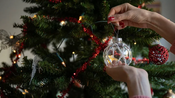 在装饰过的圣诞树上挂着光彩夺目的透明而白色的节日饰物的女性手的布景 — 图库照片