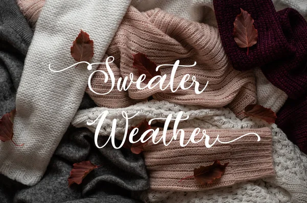 Background with warm sweaters and the inscription SWEATER WEATHER. Pile of knitted clothes with autumn leaves, warm background, knitwear, Autumn concept.