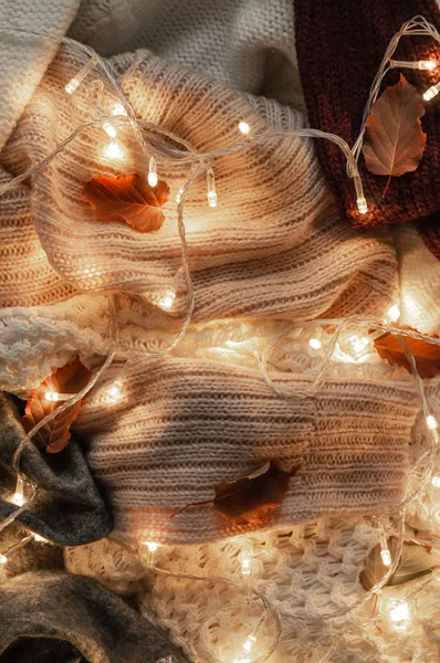 Background with warm sweaters. Pile of knitted clothes with autumn leaves and a garland, warm background, knitwear, space for text, Autumn winter concept. Copy Space.