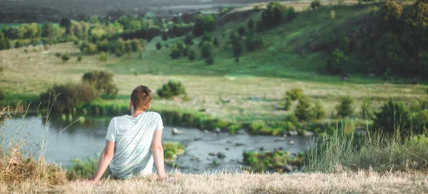 ชายหนุ่มนั่งบนเนินเขาในหญ้าและเพลิดเพลินกับภูมิทัศน์อันงดงามของธรรมชาติและเพลิดเพลินกับวิว ความสําเร็จของผู้ชนะ — ภาพถ่ายสต็อก