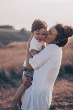 Genç Anne küçük oğlu, kisses günbatımında çayırda güneşli. Oğlu tutan anne. Anneler günü. Aile kavramı