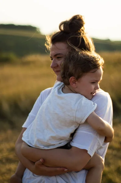 Giovane madre bacia il suo piccolo figlio al sole nel prato al tramonto. Madre che tiene in braccio suo figlio. Festa della Mamma. Concetto di famiglia — Foto Stock