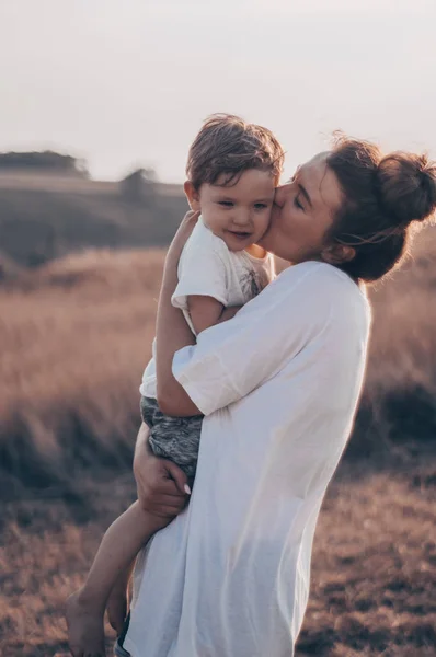 Giovane madre bacia il suo piccolo figlio al sole nel prato al tramonto. Madre che tiene in braccio suo figlio. Festa della Mamma. Concetto di famiglia — Foto Stock