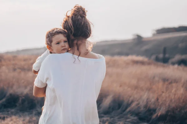 Giovane madre bacia il suo piccolo figlio al sole nel prato al tramonto. Madre che tiene in braccio suo figlio. Festa della Mamma. Concetto di famiglia — Foto Stock