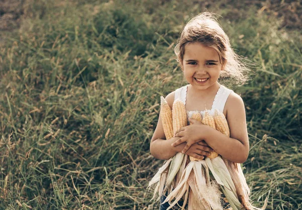 Kleines Mädchen, das im Herbst in einem Maisfeld spielt. Kind mit einem Maiskolben in der Hand. Ernten mit Kindern. Herbstaktivitäten für Kinder — Stockfoto