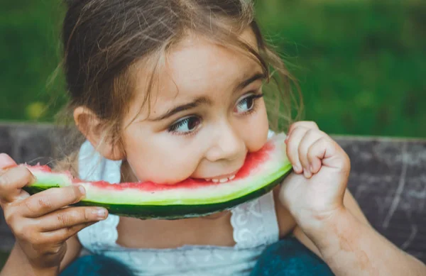 Ребёнок ест арбуз в саду. Дети едят фрукты на открытом воздухе. Здоровые закуски для детей. Красивый фон, эмоциональная девушка — стоковое фото