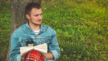 Genç adam arka plan sonbaharda tatiller ve yaşam tarzı kavramı üzerinde açık bir sıcak kırmızı kareli ve bir fincan çay ile kitap okuma gülümseyerek