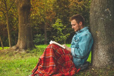 Genç adam arka plan sonbaharda tatiller ve yaşam tarzı kavramı üzerinde açık bir sıcak kırmızı kareli ve bir fincan çay ile kitap okuma gülümseyerek