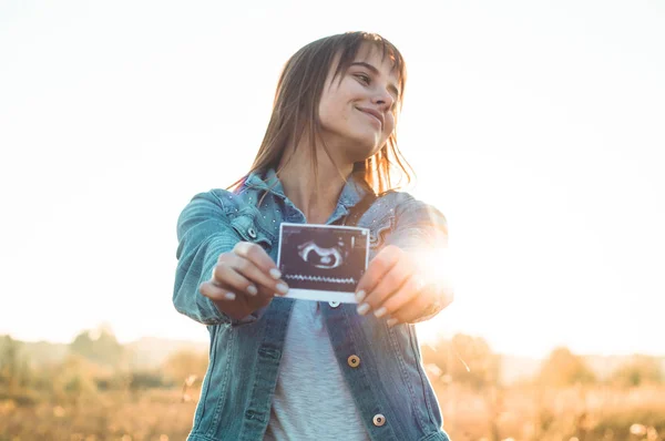 Young Pregnant Woman holding ultrasound photo at Sunset and Embracing her Belly. 4 Month Pregnancy. Maternity Concept. Toned Photo — Stock Photo, Image