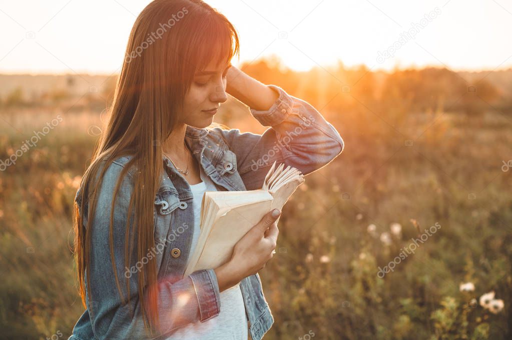 Beautiful girl in autumn field reading a book. The girl sitting on a grass, reading a book. Rest and reading. Outdoor reading