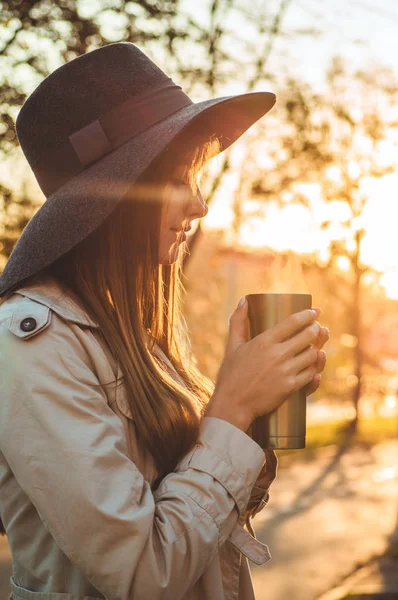 Jeune femme dans un chapeau avec thermos thermo tasse portrait extérieur en lumière du jour douce et ensoleillée. Automne. Coucher de soleil. Confortable — Photo