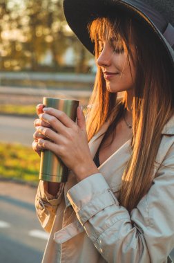 Şapkalı bir genç kadınla termos termo Kupası açık portre yumuşak güneşli gün ışığında. Sonbahar. Gün batımı. Rahat.