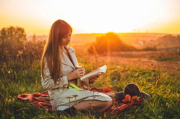 Het concept van levensstijl outdoor recreatie in het najaar. Meisje met hoed lezen boeken op plaid met een thermo-kop. Herfst. Zonsondergang. Gezellige — Stockfoto