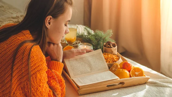 Douce photo confortable de la femme en pull orange chaud sur le lit avec une tasse de thé et de fruits. Fille assise sur le lit avec de vieux livres . — Photo