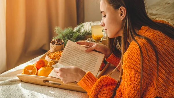 Suave foto acogedora de la mujer en suéter naranja cálido en la cama con taza de té y fruta. Chica sentada en la cama con libros viejos . — Foto de Stock