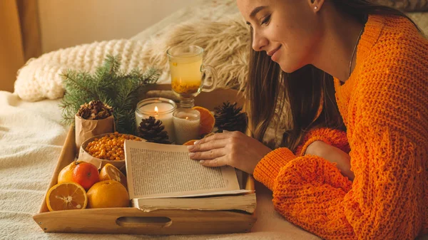 Suave foto acogedora de la mujer en suéter naranja cálido en la cama con taza de té y fruta. Chica sentada en la cama con libros viejos . — Foto de Stock