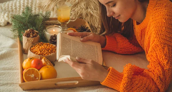 Foto aconchegante suave de mulher em suéter laranja quente na cama com xícara de chá e frutas. Menina sentada na cama com livros antigos . — Fotografia de Stock
