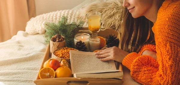 Foto aconchegante suave de mulher em suéter laranja quente na cama com xícara de chá e frutas. Menina sentada na cama com livros antigos . — Fotografia de Stock