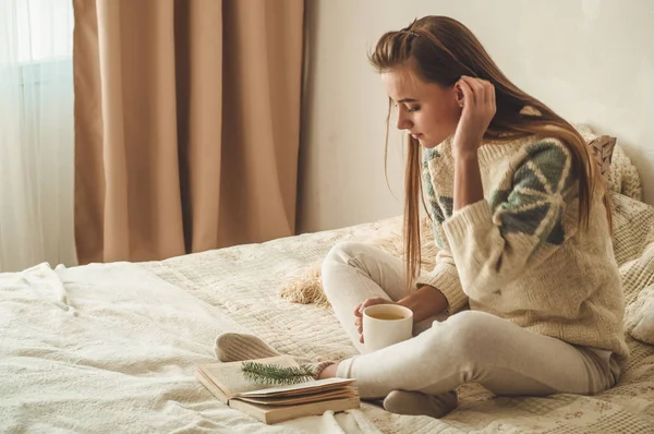 Acogedora casa. Hermosa chica está leyendo un libro en la cama. Buenos días con el té. Una chica joven y bonita relajándose. El concepto de lectura — Foto de Stock