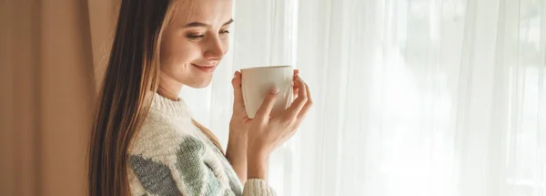 Уютный дом. Женщина с чашкой горячего напитка у окна. Смотрю в окно и пью чай. Доброе утро с чаем. Девочка расслабляется — стоковое фото