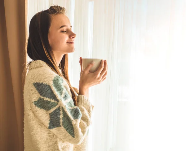 Uma casa acolhedora. Mulher com xícara de bebida quente pela janela. Olhando para a janela e beber chá. Bom dia com o chá. Menina jovem relaxante — Fotografia de Stock