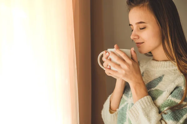 Ζεστό, άνετο σπίτι. Γυναίκα με φλιτζάνι ζεστό ρόφημα από το παράθυρο. Κοιτάζοντας από το παράθυρο και να πιείτε καφέ. Καλημέρα με τσάι. Νεαρό κορίτσι χαλάρωση — Φωτογραφία Αρχείου