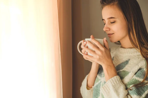 Acogedora casa. Mujer con una taza de bebida caliente junto a la ventana. Mirando la ventana y bebiendo té. Buenos días con el té. Chica joven relajante — Foto de Stock