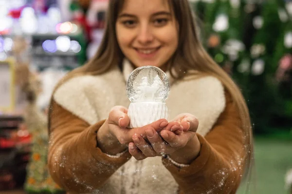 Jente i varm frakk som holder glasskule med firtrær, hus og kunstig snø på et kjøpesenter på julemarkedet. Vinterstemning – stockfoto