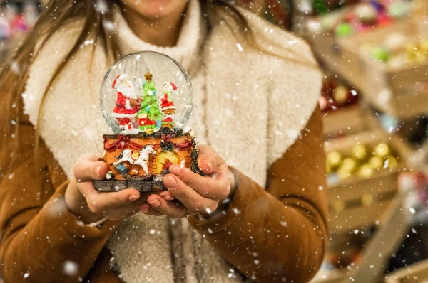 Jente i varm frakk som holder glasskule med firtrær, hus og kunstig snø på et kjøpesenter på julemarkedet. Vinterstemning – stockfoto