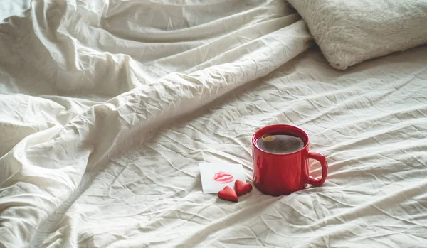 Taza roja de té y dos galletas de corazones, una nota con un beso en una cama blanca. Concepto de San Valentín — Foto de Stock