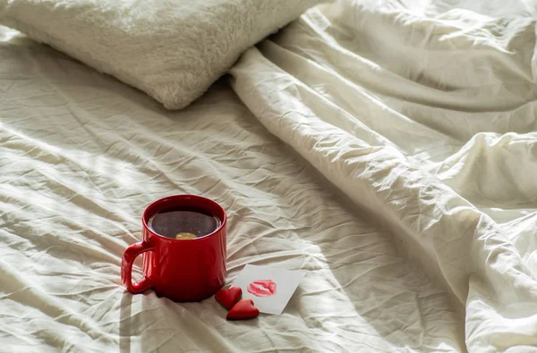 Kırmızı fincan çay ve iki kalp kurabiye, bir öpücük beyaz bir yatak üzerinde bir notla. Sevgililer günü kavramı — Stok fotoğraf