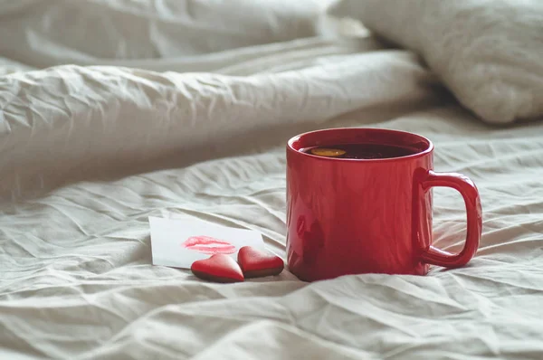 Cangkir teh merah dan dua kue hati, catatan dengan ciuman di tempat tidur putih. Valentines konsep hari — Stok Foto