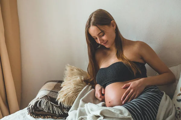 Mulher grávida atraente está sentada na cama e segurando sua barriga. Últimos meses de gravidez. — Fotografia de Stock