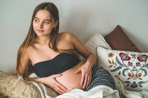 Ελκυστική έγκυος γυναίκα κάθεται στο κρεβάτι και κρατά την κοιλιά της. Τελευταίοι μήνες της εγκυμοσύνης. — Φωτογραφία Αρχείου