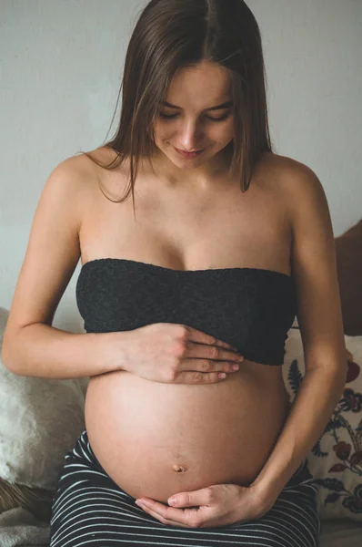 Atractiva mujer embarazada está sentada en la cama y sosteniendo su vientre. Últimos meses de embarazo. — Foto de Stock