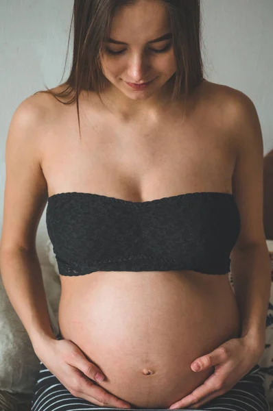 Atractiva mujer embarazada está sentada en la cama y sosteniendo su vientre. Últimos meses de embarazo. — Foto de Stock