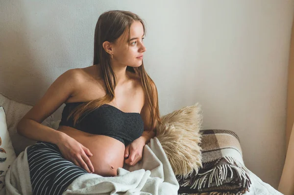 Jolie femme enceinte est assise dans son lit et tenant son ventre. Derniers mois de grossesse. — Photo