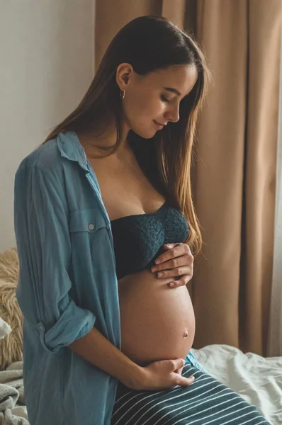 Mulher grávida atraente está sentada na cama e segurando sua barriga. Últimos meses de gravidez. — Fotografia de Stock