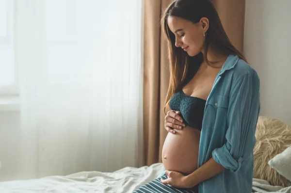 Atractiva mujer embarazada está sentada en la cama y sosteniendo su vientre. Últimos meses de embarazo. — Foto de Stock