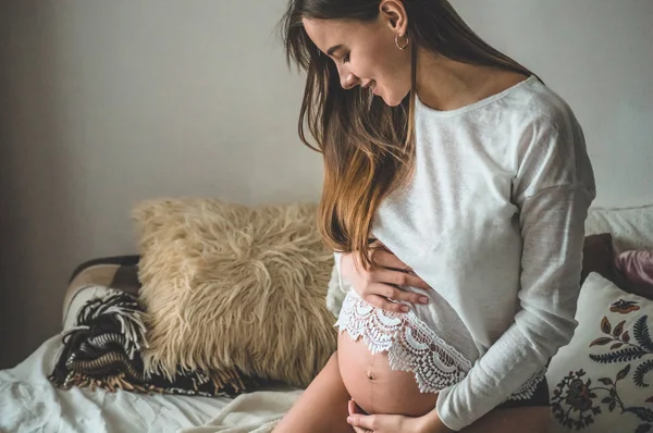 Atrakcyjna ciężarna kobieta siedzi w łóżku i trzyma brzuch. Ostatnie miesiące ciąży. — Zdjęcie stockowe