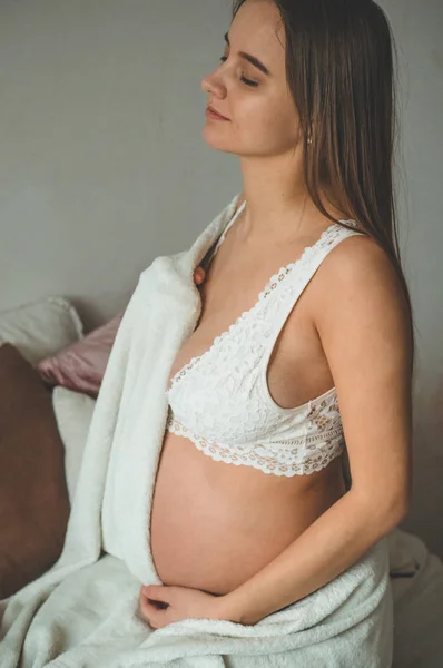 Mulher grávida atraente está sentada na cama e segurando sua barriga. Últimos meses de gravidez. — Fotografia de Stock