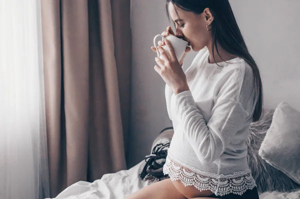 Aantrekkelijke zwangere vrouw drinkt thee op het bed. Het drinken van thee op zoek via een raam bij u thuis. Laatste maanden van de zwangerschap. — Stockfoto