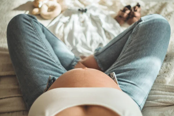 아름 다운 임신한 여자는 침대에 앉아서는 kidswear에서 보인다. 기대입니다. 임신의 개념 — 스톡 사진
