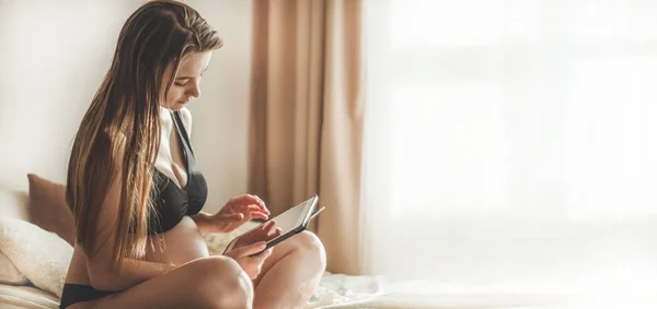 Wanita hamil dengan tablet modern duduk di tempat tidur dan menyesuaikan rambut panjangnya. konsep kehamilan dan teknologi — Stok Foto