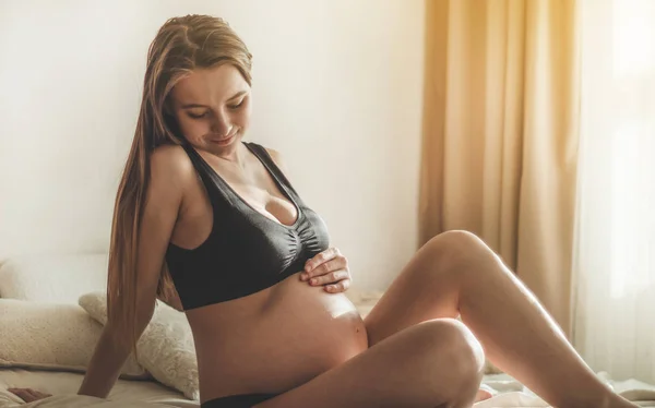 Attraktive schwangere Frau sitzt im Bett und hält ihren Bauch. — Stockfoto
