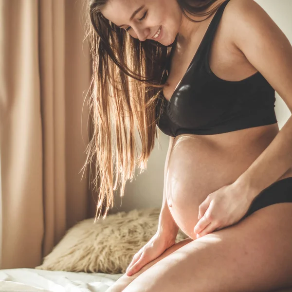 Atractiva mujer embarazada está sentada en la cama y sosteniendo su vientre . — Foto de Stock