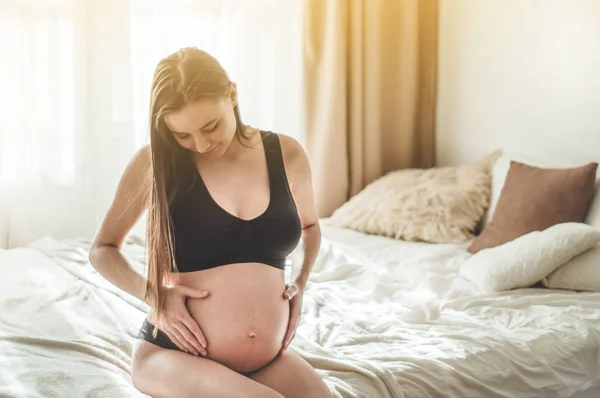 Attraktive schwangere Frau sitzt im Bett und hält ihren Bauch. — Stockfoto