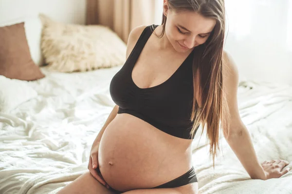 Atractiva mujer embarazada está sentada en la cama y sosteniendo su vientre . — Foto de Stock