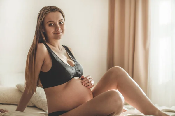 Ελκυστική γυναίκα έγκυος κάθεται στο κρεβάτι και κρατώντας την κοιλιά. — Φωτογραφία Αρχείου