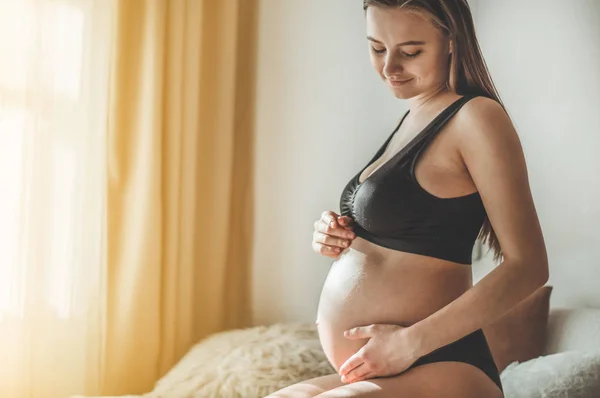 Attraktive schwangere Frau sitzt im Bett und hält ihren Bauch. — Stockfoto